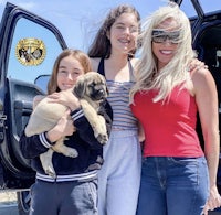 three women standing in front of a car holding a puppy