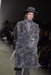 a model walks down the runway in a grey coat and hat