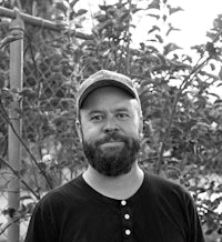 a black and white photo of a bearded man standing in front of a tree