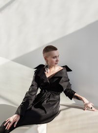 a woman in a black trench coat sitting on a white wall