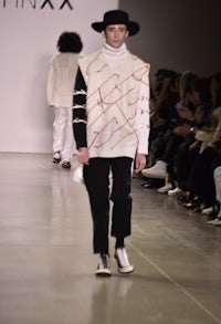 a man walks down the runway wearing a hat and a sweater