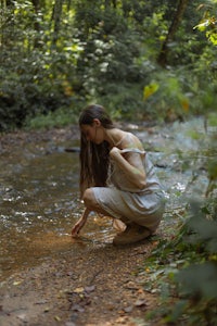 a woman crouching down next to a stream in the woods