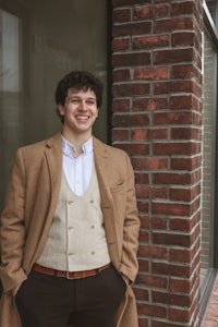 a young man in a tan coat standing in front of a brick building