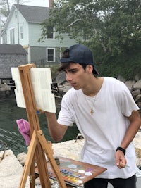 a young man painting on an easel in front of a body of water