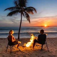 two people sitting around a fire on the beach at sunset