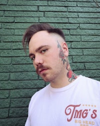 a man with tattoos and a t - shirt in front of a brick wall