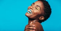 “African American woman with vibrant blue lipstick and yellow eyeliner, laughing joyfully against a bright blue background. Her short curly hair and radiant smile convey confidence and happiness. This image captures the essence of bold makeup artistry and self-expression.”