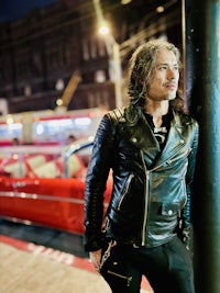 a man in a leather jacket leaning against a pole