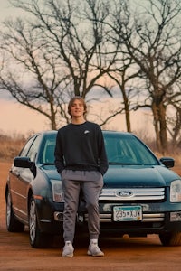 a young man standing next to his car in a dirt field