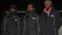 three men in black jackets standing next to a car at night