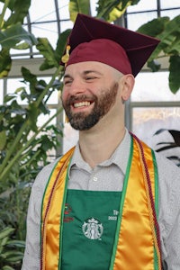 a man in a graduation cap and gown smiles in front of a plant