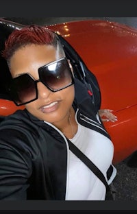a woman in a black shirt and sunglasses is posing in front of a red car