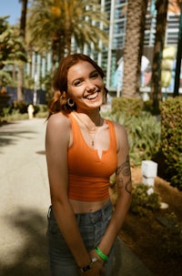 a woman in an orange top and jeans smiles in front of palm trees