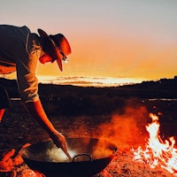 a man cooking over a campfire at sunset