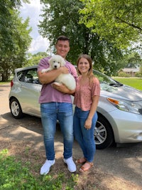 a man and woman standing in front of a silver car with a puppy