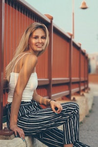a young woman wearing striped pants and a white top leaning against a wall