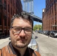 a man wearing glasses and a leather jacket in front of the brooklyn bridge