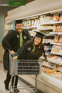 a couple in graduation gowns pushing a shopping cart in a grocery store