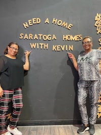 two women standing in front of a sign that says need a home saragoga homes with kevin