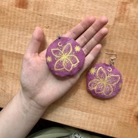 a hand holding a pair of purple flower earrings