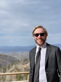 a man in a suit and sunglasses smiling in front of a mountain