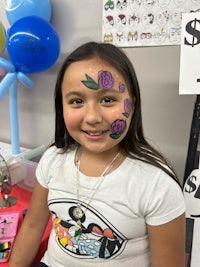 a young girl with a flower face paint smiles in front of a table
