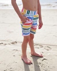 a young boy standing on the beach wearing colorful striped shorts
