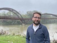 a man with glasses standing in front of a bridge