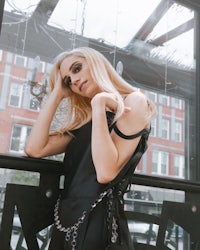 a blonde woman in a black dress leaning against a window