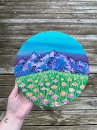 a person holding up a painting of mountains and flowers