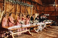 a group of bridesmaids and groomsmen standing at a long table in a barn