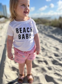 a little girl standing on the beach wearing a beach babe t - shirt