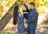 a family is holding their daughter in front of a tree in the fall
