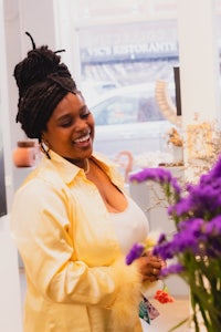 a woman with dreadlocks smiles in front of a vase of purple flowers