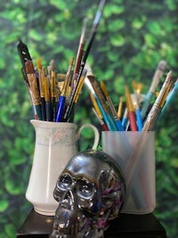 a metal skull sits on a table next to paint brushes and other art supplies