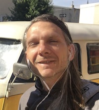 a man with long hair standing in front of a yellow vw bus