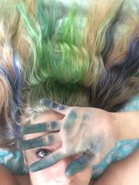 a woman with green and blue hair covering her face with her hands