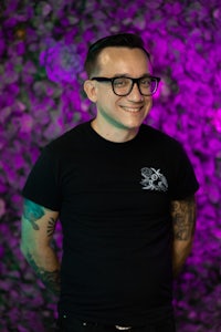 a man with glasses and tattoos standing in front of a purple wall