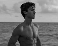 a black and white photo of a shirtless man standing by the ocean