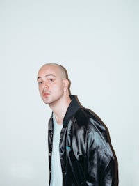 a bald man in a black jacket standing in front of a white wall