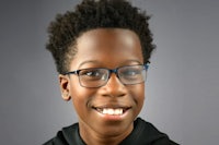 a young boy wearing glasses smiles in front of a gray background